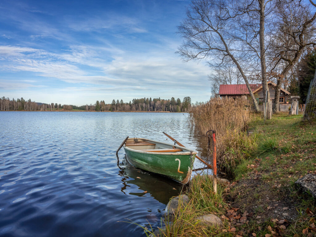 Am Seeufer ist ein Ruderboot verankert. Am Ufer hinter einem Baum ist das Fischerhäusl zu sehen.