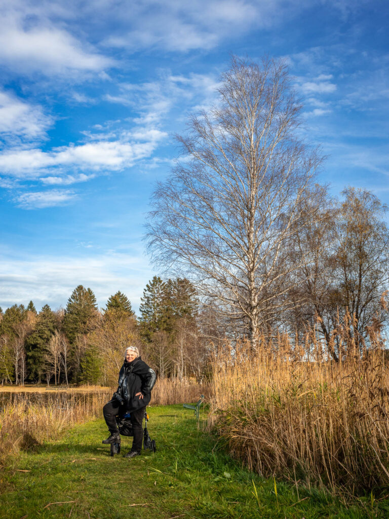 Meine Frau Diana steht mir ihrem Rollator auf einem Grünstreifen am Seeufer vom Bayersoiener See neben einem Schilfstreifen und einer Birke. Im Hintergrund ein Mischwald.