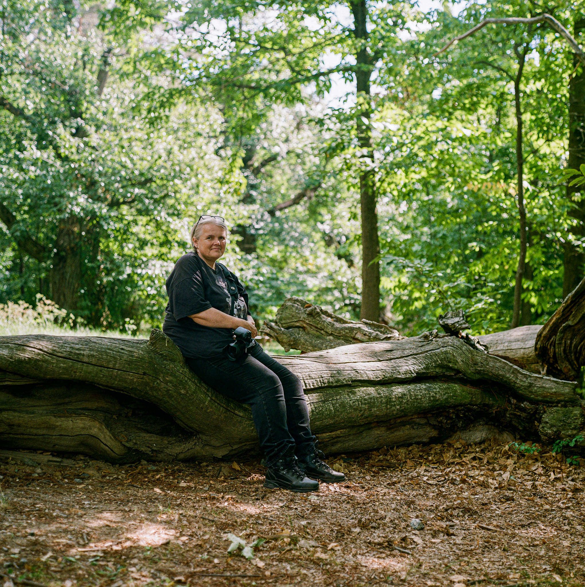 Auf dem Farbfoto sitzt meine Frau zwischen Kastanienbäumen auf einem umgefallenen Baumstamm und lächelt in die Kamera.