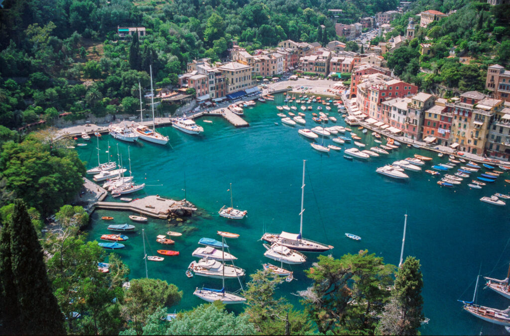 Ein Farbfoto aufgenommen von einem erhöhtem Standort mit Blick auf die Bucht von Portofino. Im tief grünblauen Meer liegen viele Jachten und an der Bucht stehen die bunten Häuser des Ortes. bunten Häuser, das grünblaue Meer und auf vielen Jachten.