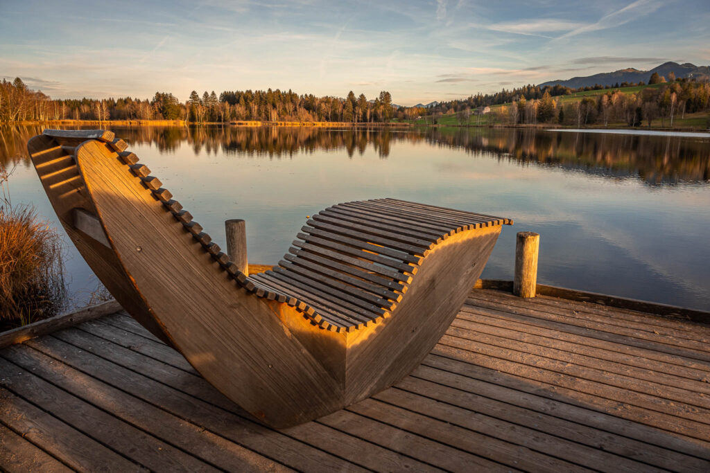 Ein Steg mit einer schönen, hölzernen, ergonomisch geformten Liege im warmen Licht der Abendsonne. Hinter dem Bayersoiener See erstrecken sich ein Wald und einige Berge.